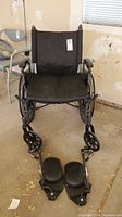 Front view of the black wheelchair showing seat, backrest, armrests, and detached footrests on the floor.