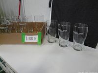 12 Barley Days beer glasses, 9 inside a cardboard box and 3 outside, shown clearly with visible branding and shape.