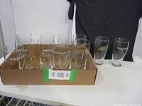 Box holding 12 clear beer glasses with Barley Days Brewery logos, arranged in rows, displayed on a wooden surface with a dark t-shirt in background.