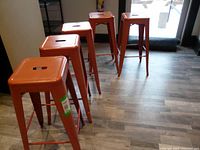 Five metal stools arranged standing individually showing terra cotta color and height on a wood-look floor.