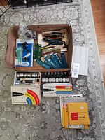 Top-down view of cardboard box containing drafting pencils, boxes of mechanical lead refills, drafting inks, film papers, and accessories on carpeted floor.