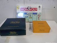 Photo showing sealed Monopoly game at the back with two Trivial Pursuit boxes in front, blue and orange, all placed on a white surface against a white background.