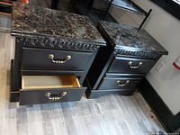 Two black night tables with faux marble tops side by side, one with a drawer partially open to show interior.
