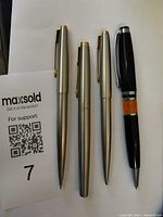Photo showing four writing instruments together: three silver-tone (two pens, one pencil) and one black Franklin Covey pen with amber accent.