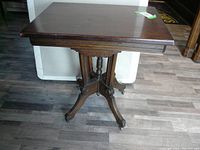 Full view of vintage Eastlake style side table showing square top and carved base with casters on wood flooring.
