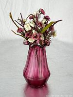 Full view of cranberry glass vase with faux floral arrangement placed inside, showing color and shape of vase and flower types.