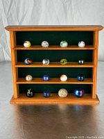 Front view of wooden display case filled with 16 large marbles arranged on four shelves with green felt background.