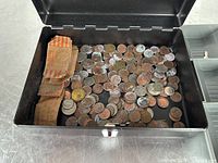 Open cash box showing loose change including wrapped coin rolls and various coins in the main compartment and a removable plastic tray.