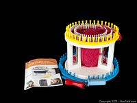 KB rotating double knit loom with red yarn wound around center, yellow and blue removable knit rings, two plastic knitting hooks (red and blue), instruction booklet on black background.