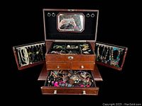 Open jewelry box showing interior compartments with assorted jewelry, including necklaces and earrings on side panels, and bracelets, rings, and other pieces in drawers.
