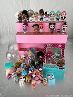 Front view showing the 18 LOL Surprise mini dolls arranged on a pink multi-tiered display stand with various accessories below and a boxed accessory set.