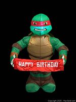 Front view of the green inflatable Raphael character holding the red Happy Birthday sign with visible facial expression and shell details.