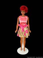 Full-length image of vintage 1960s Mattel Barbie Julia doll standing on a white stand, wearing original pink satin and gold floral dress with pink sash tied at the waist.