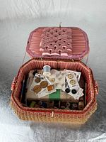 Open view of vintage woven sewing basket showing buttons on cards, assorted wooden and plastic thread spools inside, and pink satin pin cushion attached to the lid interior.