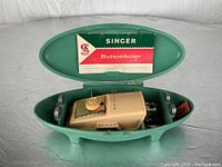 Top-down view of vintage Singer buttonholer secured inside green oval case with instruction booklet inside lid.