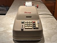 Front angle view of antique Monroe adding machine plugged in, showing numeric keypad, plus key, and paper roll at back.