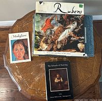 Photo showing four art books including Rubens, Modigliani, Louvre, and Ten Schools of Painting on a wooden octagonal table.
