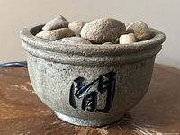 Close-up image of the Asian style ceramic bowl water fountain topped with smooth natural rocks and showing black Asian characters on the side.
