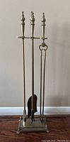 Full view of the brass fireplace tool set including stand, poker, shovel, and tongs placed on hardwood floor against white wall.