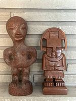 Two carved Mexican figurines placed side by side in front of a gray stone wall background. The ceramic figurine is on the left, reddish-brown in color with abstract features. The wooden figurine is on the right, brown with detailed carvings and visible cracking.