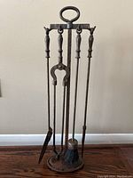 Cast iron fireplace tool set with four tools on a stand against a beige wall on wood flooring.