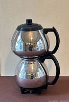 Front view of stainless steel electric coffee percolator showing stacked chambers, black handles, and lid.