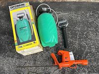 Optimum 3-gallon manual sprayer next to its original box, Black and Decker 16-inch hedge trimmer, and sprayer pump handle on asphalt surface.
