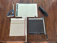 Top-down view of lot showing two guillotine paper cutters with grid cutting surfaces, white paper trimmer, and metal protractor set square on wooden table