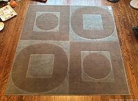 Top-down photo of square carpet on wooden floor. Carpet features four quadrants with geometric shapes - two with circles inside squares, two with squares inside circles, in muted grey and brown tones.