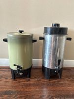 Two coffee percolators side-by-side, one green and one metallic silver, showing front spouts and side handles on a hardwood floor.