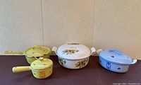 Front view of four vintage enamel cookware pieces showing yellow small pots, cream floral casserole, blue oval casserole.