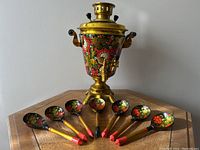 Full view of the samovar and seven hand-painted wooden spoons with floral designs on wooden surface.