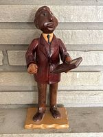 Front view of the wooden carved figurine of a boy dressed in formal suit holding an open book, placed against a stone wall background.