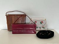 Four vintage handbags arranged on a white surface against a white wall: brown snakeskin handbag in back left, white beaded floral handbag back right, red leather handbag centered front, black embroidered pouch in front right.