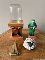 Full lot showing all four items together on a wooden surface against a light wall: wooden nut dispenser man, ceramic masked dentist figure, ceramic sheep, and brass sailboat clip.