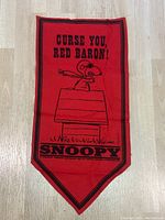 Front view of red Snoopy felt pennant featuring Snoopy flying his doghouse as the Red Baron with text 'CURSE YOU, RED BARON!' and 'SNOOPY' below.