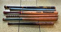 Front view of six vintage wooden baseball bats laid out horizontally on a woven mat surface, showing various brands, markings and handle wrappings, highlighting lot contents.