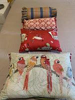 Top view of four cushions stacked: striped bolster, red damask patterned, vintage print red cushion and parrot embroidered cushion at front.
