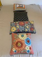 Five assorted cushions laid out on a bed showing a variety of fabrics and designs, including floral embroidery and beading, geometric and striped patterns, and solid color.