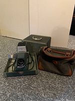 Photo showing the Argus 300 slide projector in its storage box alongside a soft camera bag, both resting on carpeted floor.