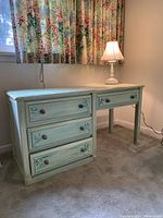 Front view of mint green desk with four drawers and decorative knobs, with table lamp switched on on top of it