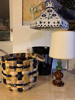 Photo showing a large woven basket with blue and tan colors, black cylindrical basket, and small desk lamp with green and brown glass base and white cylindrical lampshade.