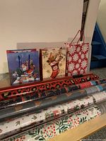Photo showing six rolls of Christmas wrapping paper, five sealed and one unsealed, along with three Christmas-themed gift bags: blue with winter scene, one with teddy bear in stocking, and red with white snowflakes.