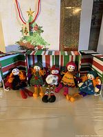 Five handmade felt doll Christmas ornaments posed sitting and standing with colorful yarn hair and festive clothing inside striped gift boxes with Christmas-themed gift bags in the background.