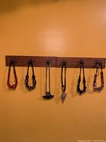 Six assorted colorful bead necklaces displayed hanging on wooden hooks against mustard yellow wall.