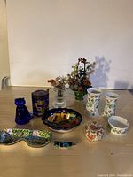 Full lot of decorative collectables on table including cobalt blue cat figurine, tin container, glass floral arrangements, candle holders and trinket dishes.