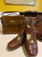 Photo showing brown Caprice Mary Jane shoes with strap, a small structured handbag with crocodile pattern and gold clasp, medium and large brown handbags.