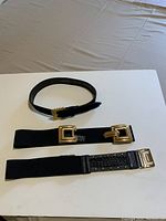 Photo showing three black ladies belts laid flat on a white surface, highlighting distinct gold color buckles and different materials (suede, vinyl, stretch).