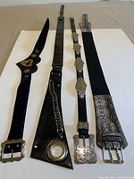 Four ladies belts laid flat on a neutral surface; inclusive of a genuine leather belt with large silver accent, two vinyl belts with ornate silver buckles, and one stretch belt with silver snake-skin design.