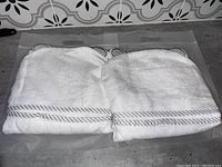 Photo showing two folded white hooded towels with grey striped trim and ear-shaped details on the hoods, placed on a grey surface.